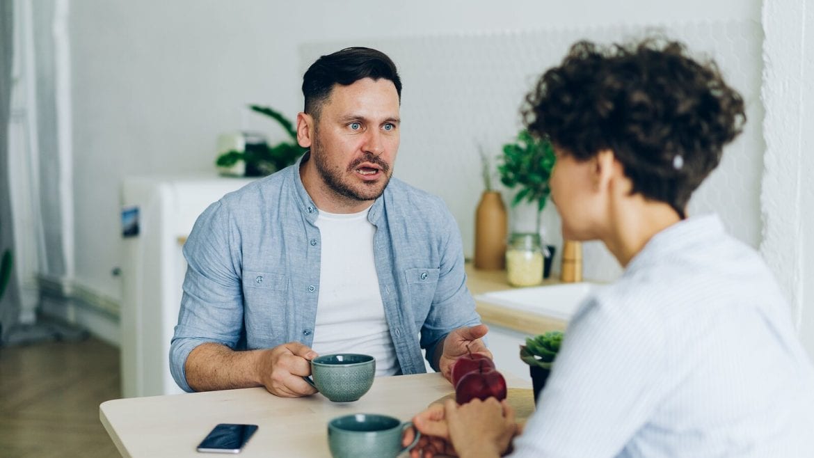 Two adults sit at a kitchen table. One appears to be talking while holding a cup, and the other listens attentively