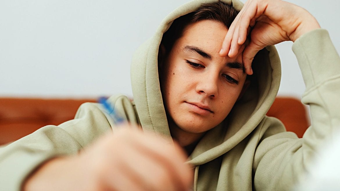 A teenager in a light green hoodie is concentrated on writing something. His head is supported by one hand, showing focus but not overt struggle