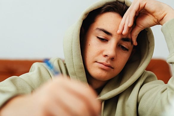 A teenager in a light green hoodie is concentrated on writing something. His head is supported by one hand, showing focus but not overt struggle