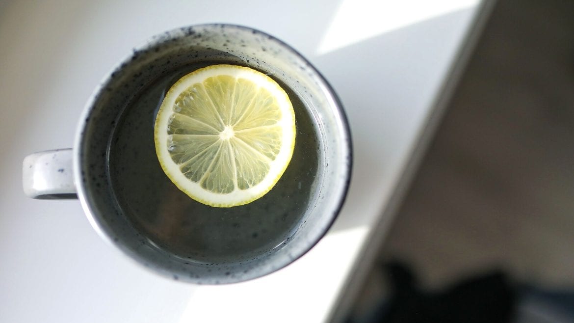 A top-down view of a cup containing water and a lemon slice. The cup is on a white surface, lit by soft, morning light