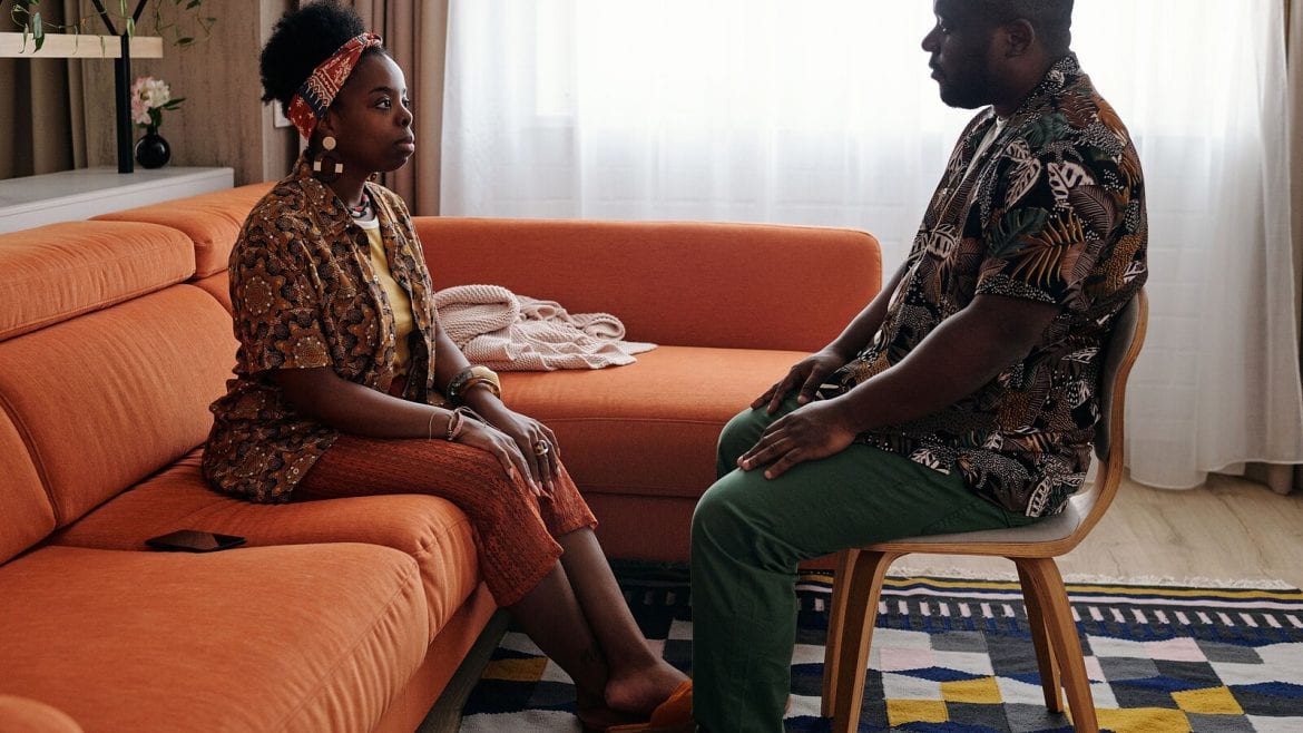 A couple sits facing each other in a brightly lit room. The woman is on a couch, looking intently at the man who is seated in a chair