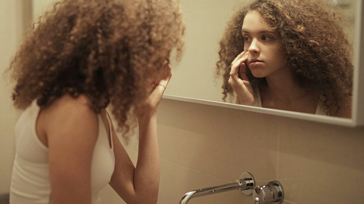 A young woman with curly hair touches her face while looking at herself in a bathroom mirror, appearing thoughtful