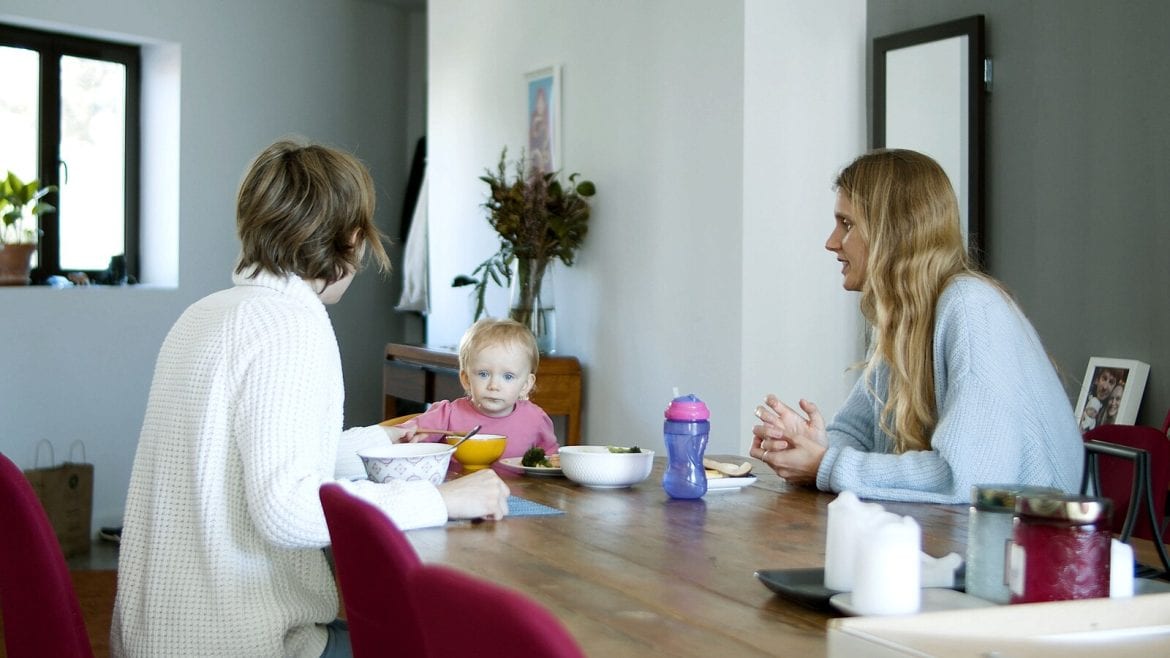 Two adults sitting at a table with a toddler, appearing to have a calm and steady conversation. The table is set with bowls and cups
