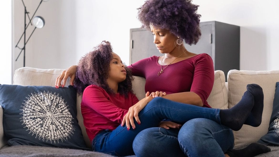 A mother is sitting on a couch with her daughter, who is leaning on her. Both appear to be having a serious conversation