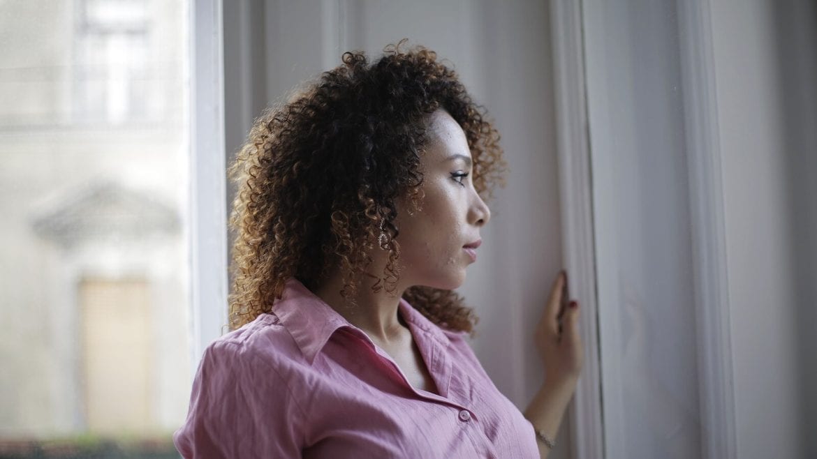 A person with curly hair is gazing out a window. They are wearing a pink shirt and appear to be in a contemplative mood