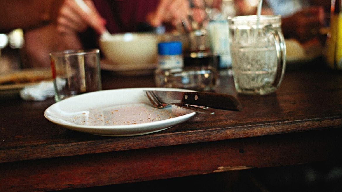 An image of an empty white dinner plate with a fork resting on it, positioned on a wooden kitchen table