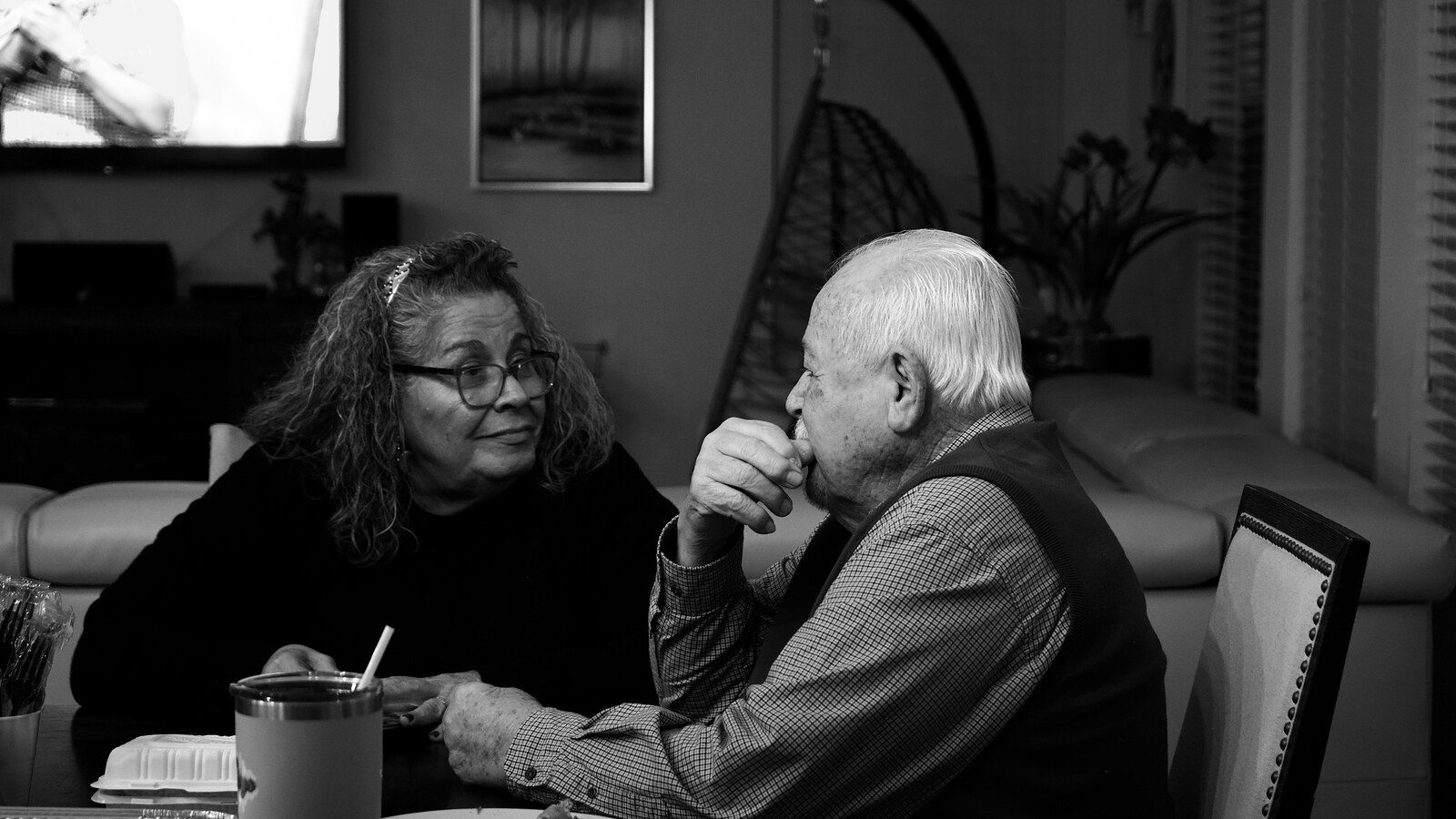 A black and white image of an older couple sitting at a table, engaged in a conversation. The woman appears to be listening intently to the man