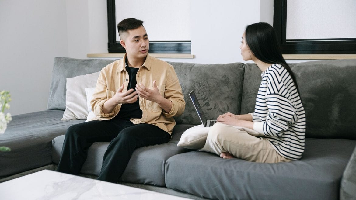Two adults are seated on a couch in a living room, engaged in conversation. One is speaking and using hand gestures, while the other is listening attentively