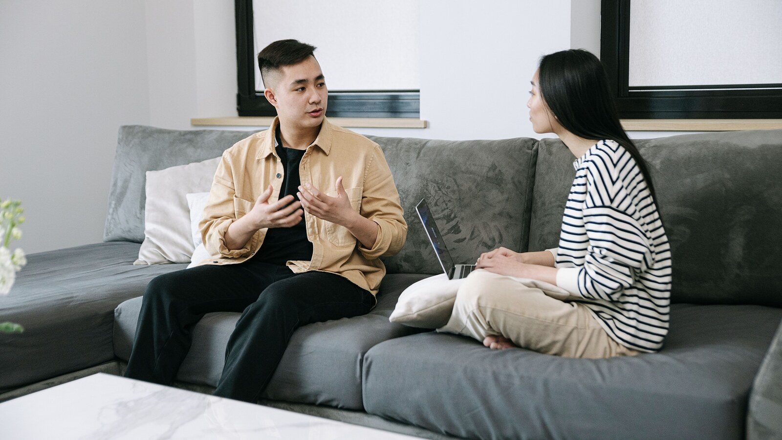 Two adults are seated on a couch in a living room, engaged in conversation. One is speaking and using hand gestures, while the other is listening attentively