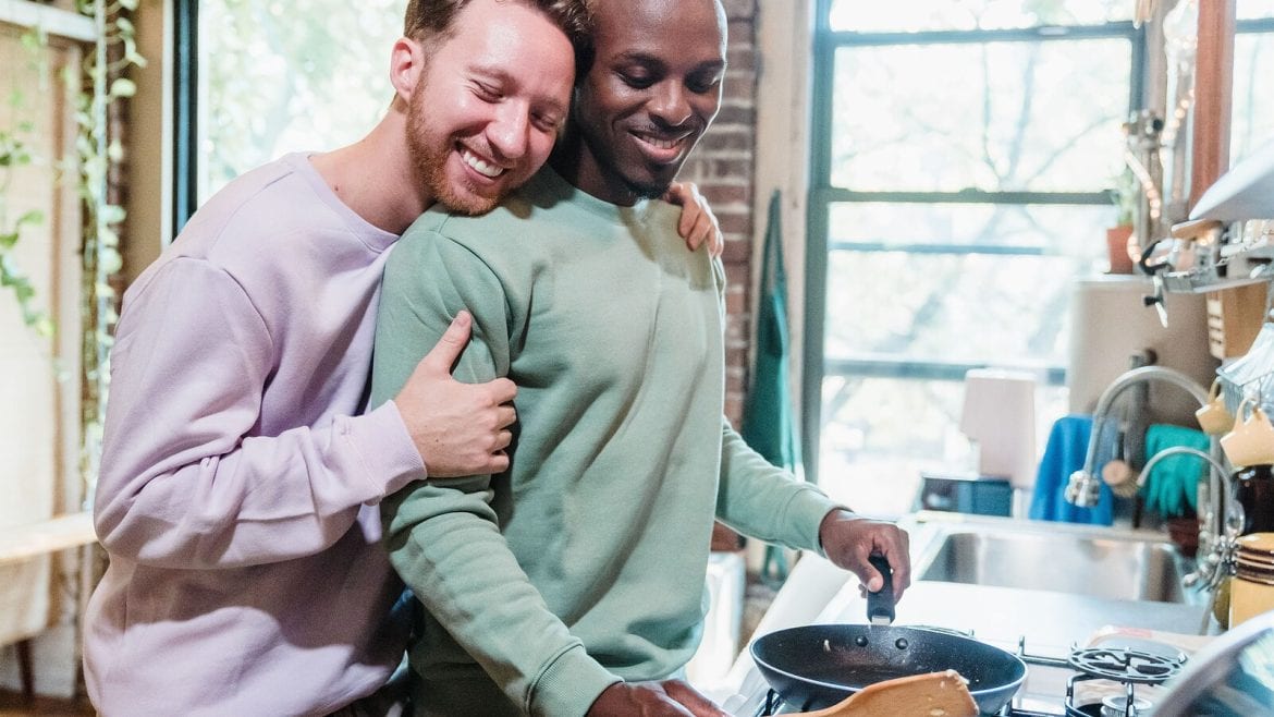 A couple is in a kitchen. One person hugs the other from behind while the second person cooks in a pan. They both appear happy and relaxed
