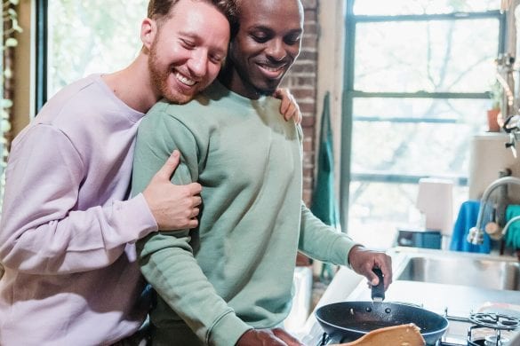 A couple is in a kitchen. One person hugs the other from behind while the second person cooks in a pan. They both appear happy and relaxed
