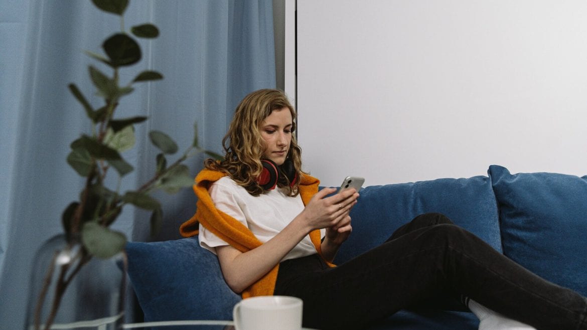 A person with headphones around their neck is sitting on a blue couch, looking at their smartphone