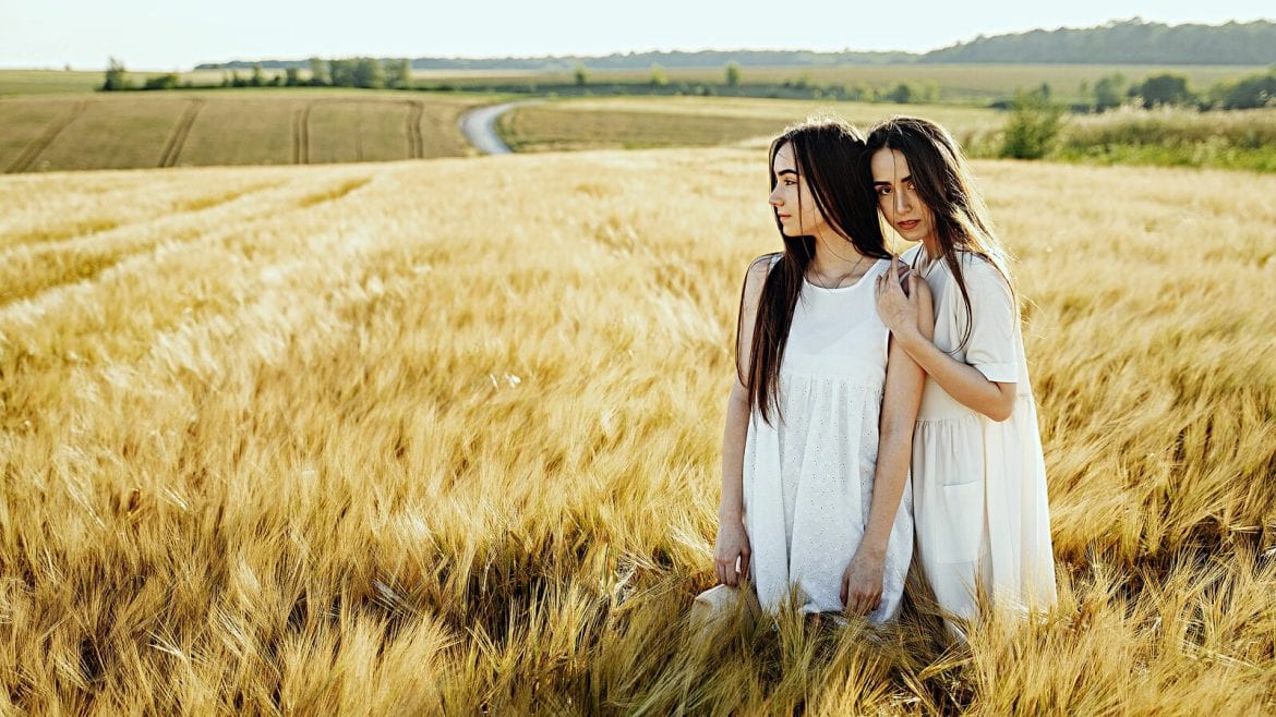 Two women standing in a wheat field, looking in different directions with a serene expression