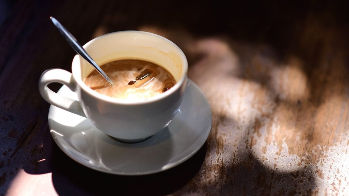 A cup of coffee with a spoon inside, placed on a wooden table under dappled sunlight creating a warm ambiance