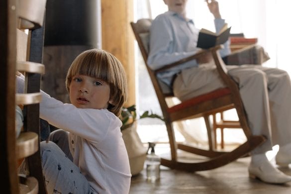 A young child sits on the floor next to a piece of furniture, while an older person is in the background seated in a rocking chair, holding a book