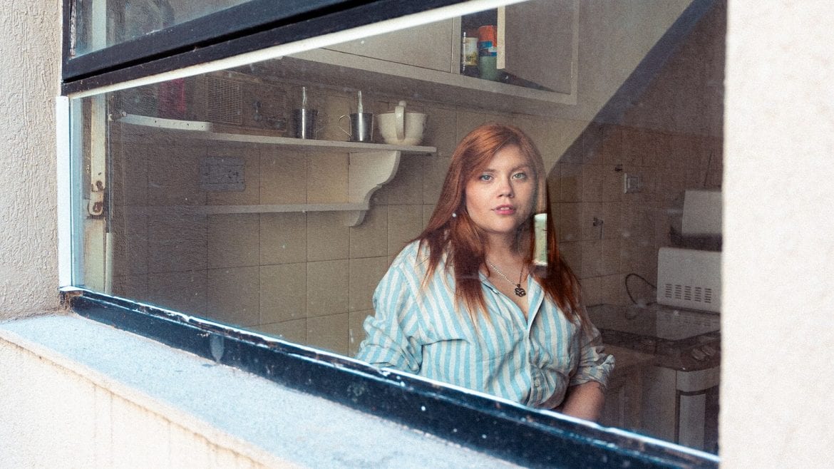 A woman with long red hair is seen through a window, wearing a striped shirt. She is standing in a kitchen, looking out as if lost in thought