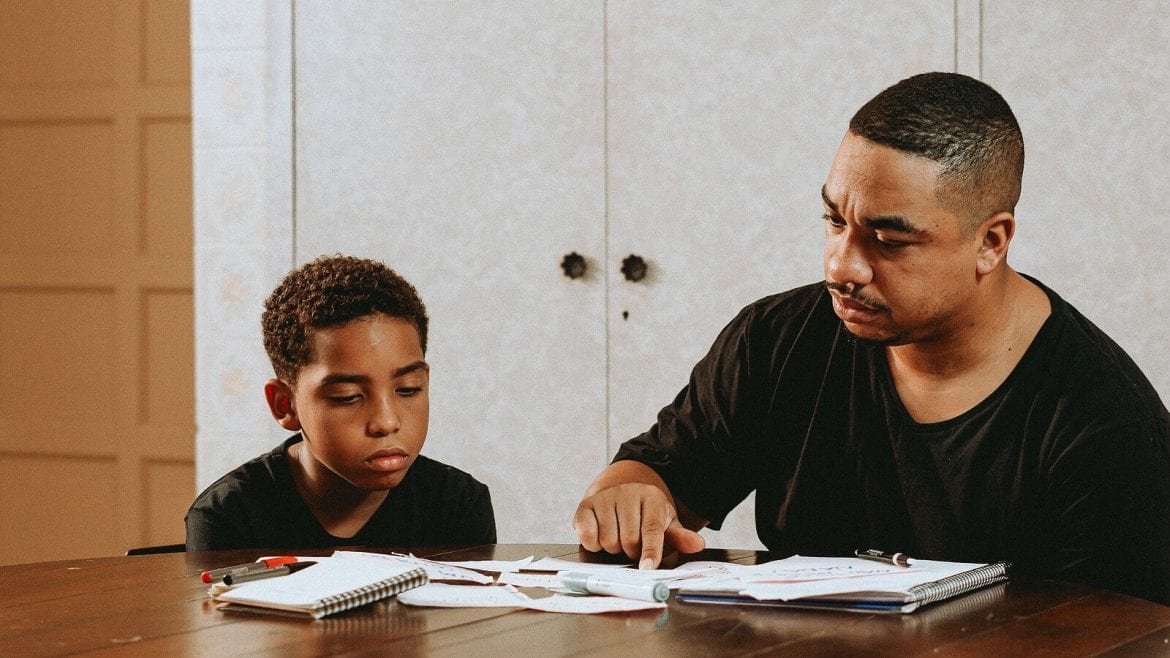 A man and a child sit at a wooden table, with papers spread out. The man is pointing at something on the papers, while the child looks at them intently