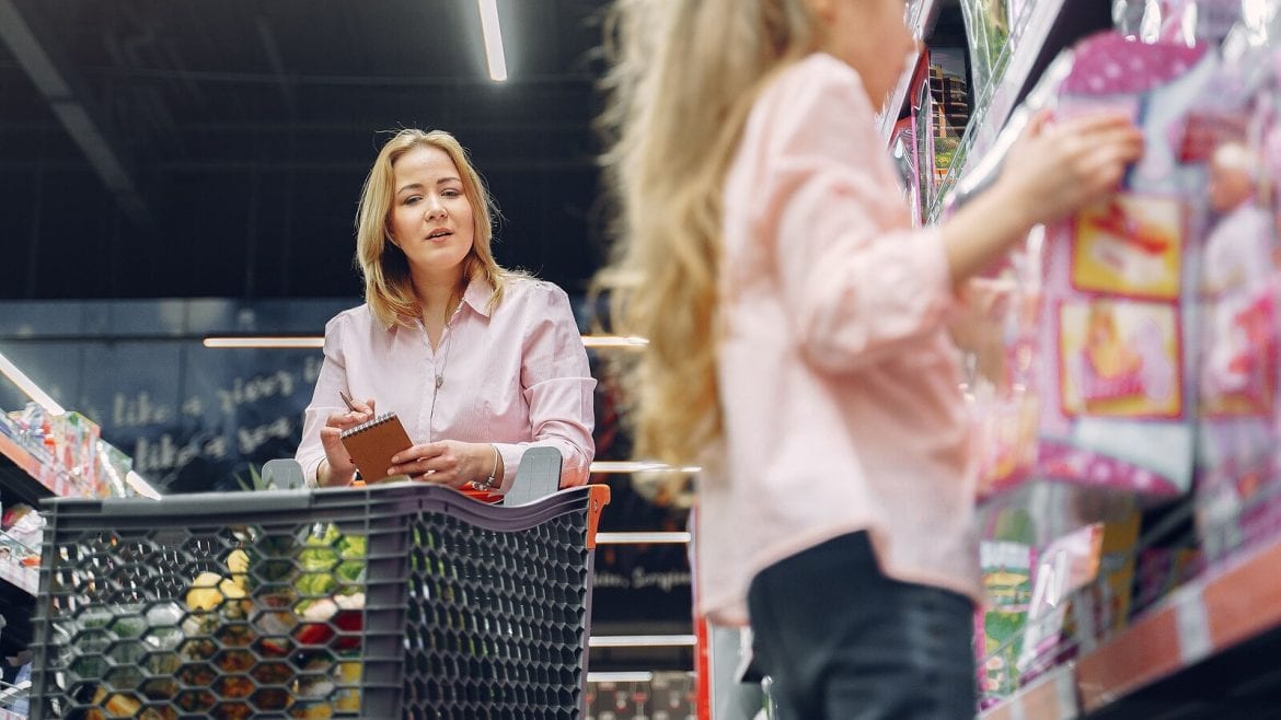 A woman is pushing a shopping cart in a grocery store, looking at a girl holding an item from the shelf