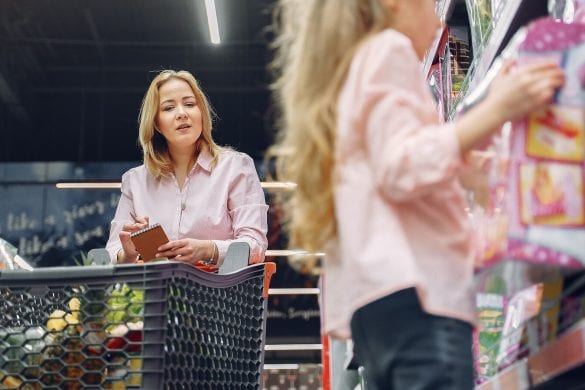 A woman is pushing a shopping cart in a grocery store, looking at a girl holding an item from the shelf