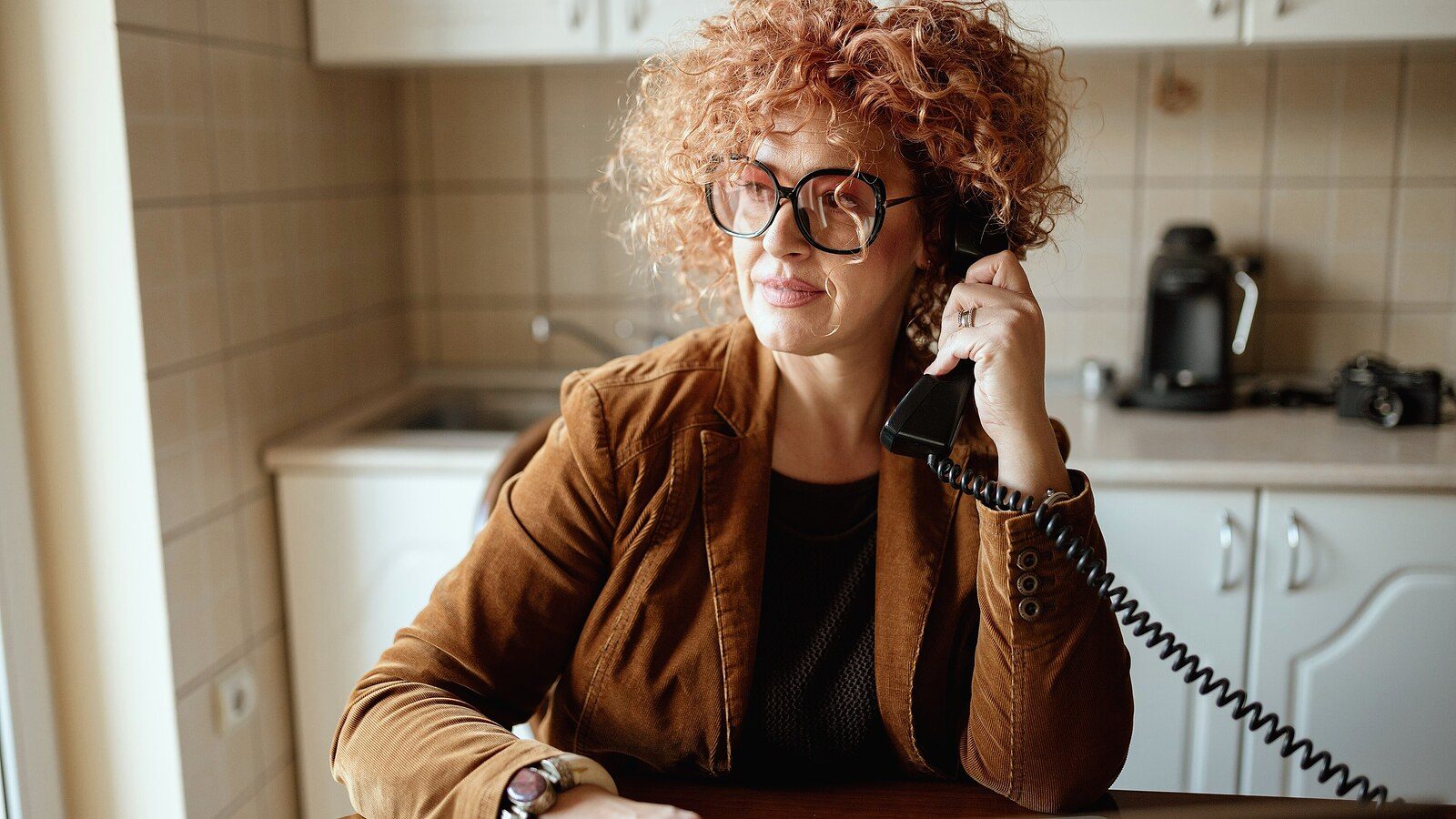 An individual with curly hair and glasses is seated at a kitchen table on the phone, looking thoughtful