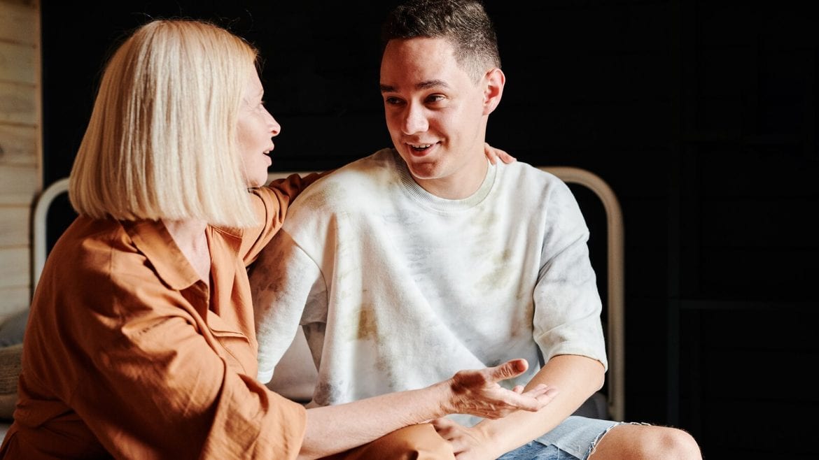 A woman with short blonde hair and a brown shirt talks to a teenage boy in a white t-shirt. They are sitting indoors, smiling and engaged in a conversation