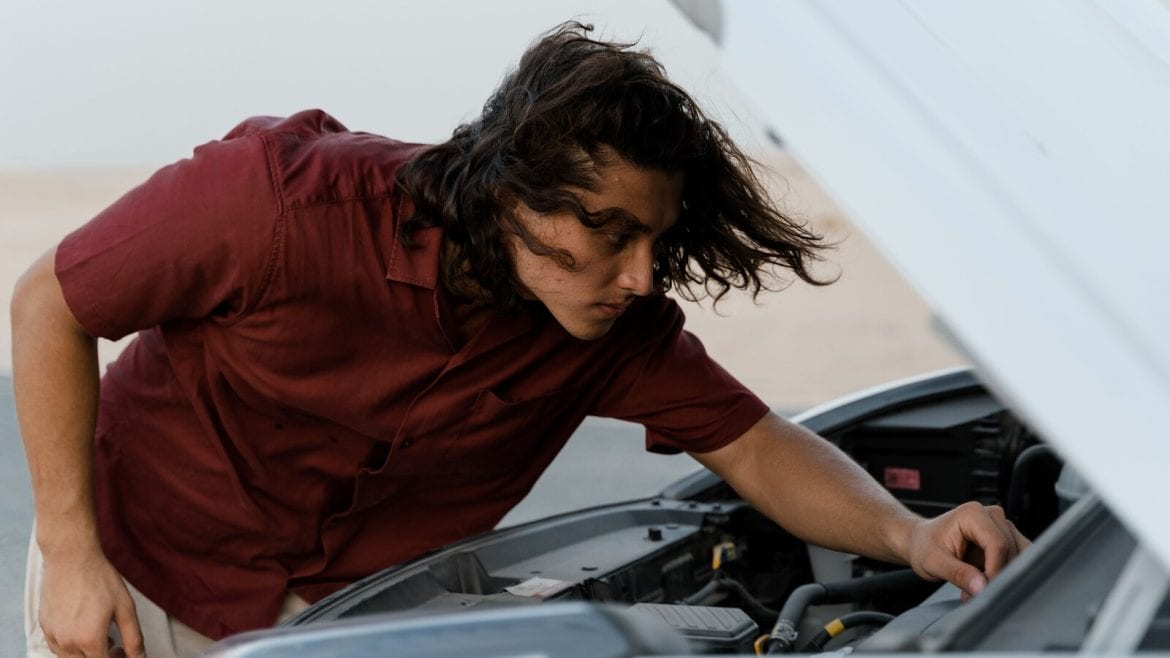 A person with long hair inspects the engine of a car with the hood opened. They are wearing a red shirt and are focused on the task