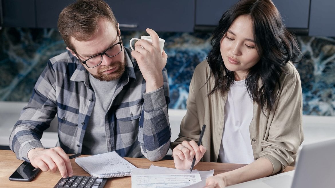 A man and woman are seated at a kitchen table. The man is using a calculator and the woman is writing something down. Papers are spread on the table